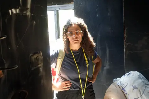 Dafna Gerstner, in glasses and with dark curly hair, stands with her hands on her hips in a burnt out house. She is wearing black trousers and a grey t-shirt and has a whistle round her neck.