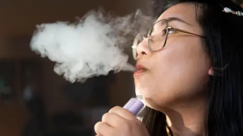 Getty Images Young woman with large metal framed glasses, vaping in a darkly lit room. There is a large cloud of smoke from her mouth.