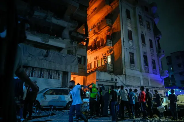 People gather in front of a damaged building after an Israeli military strike at the Nuwayri area in Beirut, Lebanon. The building glows orange, presumably from flames