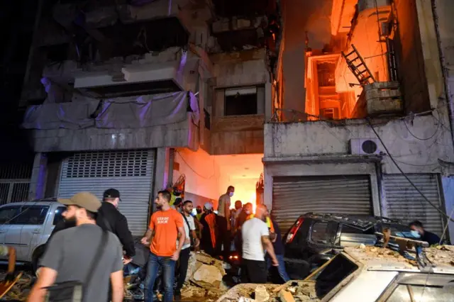 A lot of people stand on rubble in front of a burning building