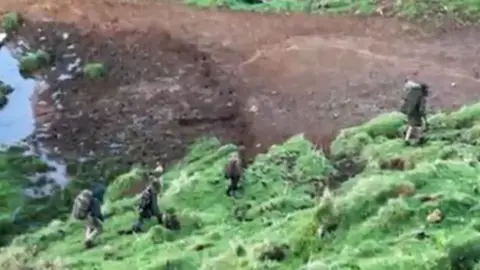 New Zealand Herald  Tom Phillips walking ahead of his three children across a bit of green ground, with darker ground visible beyond them. All three are wearing backpacks.