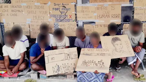 BBC Tamil migrants on Diego Garcia, with their faces blurred, holding protest signs reading "please save us" and "we need freedom", with other signs attached to a fence behind them