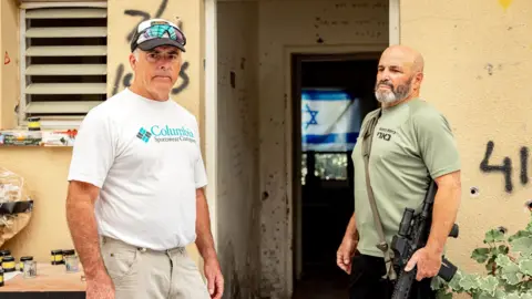 Maya Meshel / BBC Two men. One aged 60 - the other 70. One is clean-shaven wearing a white T-shirt and cap. The other has a beard, is wearing a green T-shirt and is carrying a gun