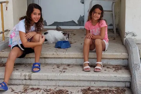 Handout Shir's daughters, both with long dark hair and smiling, sit on the steps of their home - next to them a cat eats out of a cat bowl