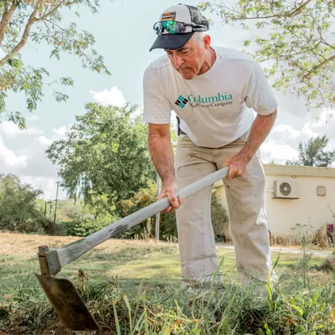 Maya Meshel / BBC Simon King, in a white T-shirt, baseball hat and beige trousers, wields a gardening tool as he tidies the kibbutz gardens. He is surrounded by greenery.