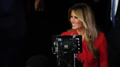 Reuters Melania Trump looks away from the camera, smiling. Video recording equipment is positioned in front of her. She is wearing a red jacket.