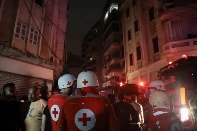 Red cross workers huddle round and look forward at a ruined building