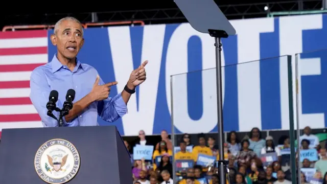 Barack Obama speaks on stage in a blue shirt, with the words 'Vote' seen in the background.