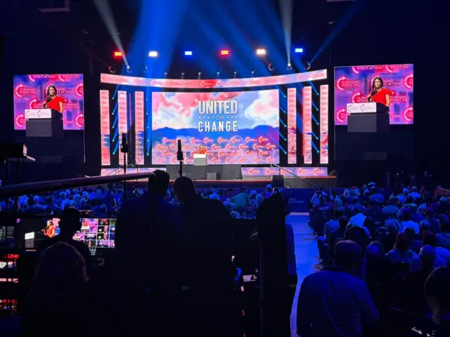 Danica Patrick appears on stage at Trump rally, the crowd is shaded blue and the screen is bright red