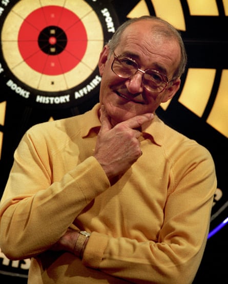 Jim Bowen poses in midshot with hand on chin with gameshow dartboard behind