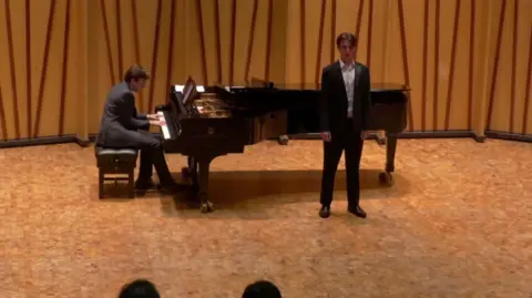 Anne Jacques Alex Harpum sings in front of an audience with his feet shoulder width apart. He is wearing a black dinner jack, black trousers and white, open-collared shirt. A pianist accompanies his singing in the background of the still picture, taken from a video of the performance. The silhouettes of the heads of members of the audience can be seen in the foreground of the image.