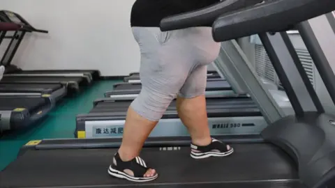 Getty Images An overweight woman wearing grey shorts and a black t-shirt walks on a treadmill