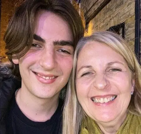 Anne Jacques Alex Harpum (left) smiles at the camera with his mouth slightly open, showing a glimpse of his teeth. His hair is centre parted and flops over his right eye slightly. He has his head leaned against that of his mother Anne Jacques who also grins with her teeth showing in this picture and squints her eyes, while wearing a green scarf like a pashmina.