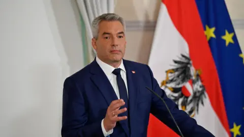 Reuters Karl Nehammer wearing a deep blue suit and a tie, standing in front of a microphone, with the Austrian and EU flags behind him