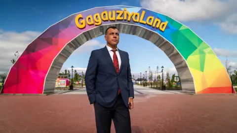 Matthew Goddard Ilya Uzun dressed in a suit, white shirt and red tie standing in front of a rainbow coloured arch that has the words GagauziyaLand written on it in yellow writing.