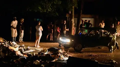 Reuters A group of people stand on a dark street in the Cuban capital Havana. Some are illuminated by the headlights of a stationary car. A pile of rubbish and rubble can be seen in the left foreground