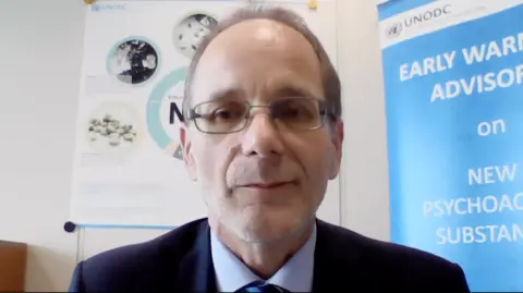 Martin Raithelhuber faces the camera while sat in an office, wearing a navy blue blazer, pastel blue shirt and navy-blue, striped tie and glasses. Behind him are two unfurled banners: one to his left is referring to an early warning advisory message from the United Nations Office on Drugs and Crime (UNODC) about new psychoactive substances (NPS); over his right shoulder the second banner describes the effects of NPS.