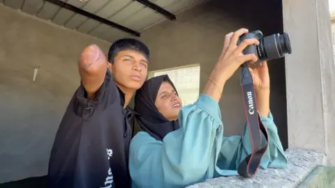 Diya al-Adini and his sister Aya stand shoulder to shoulder as Aya holds up Diya's camera taking a photo