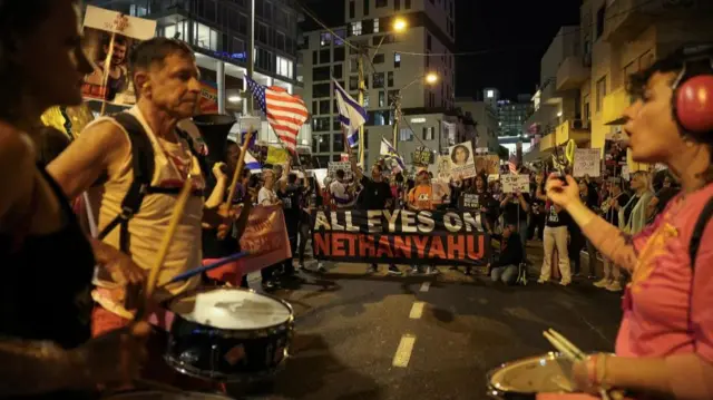 Protesters with drums line the street, others hold flags and banners, one reading 'All eyes on Netanyahu'