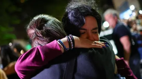 Reuters Two fans hugging outside the hotel. Facing the camera is a woman with a purple jumper on, lots of bracelets and long, dark blue nails. She has an anguished expression.