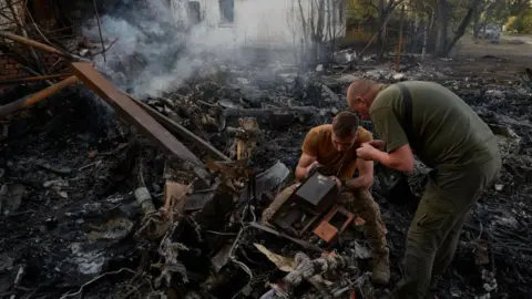 Pierre Crom/Getty Images Ukrainian servicemen examine the wreckage of a downed Russian aircraft, likely a Sukhoi S-70 "stealth" heavy unmanned combat aerial vehicle (UCAV), which crashed in a residential area, setting a house on fire on October 5, 2024 in Kostyantynivka, Ukraine