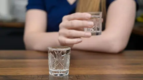 Getty Images glass containing clear liquid on a table with a woman holding a similar glass in the background