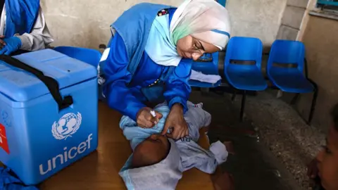 EPA A small Palestinian child lies on a table and receives oral polio vaccine drops from a healthcare worker during a vaccination campaign at UNRWA headquarters in Deir al-Balah, central Gaza Strip, 14 October 2024