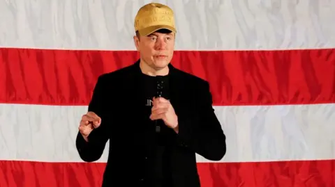 Reuters Elon Musk, wearing a gold MAGA hat, speaks into a microphone while standing in front of an American flag.