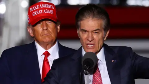 Reuters Mehmet Oz speaking into a microphone and gesturing as Donald Trump behind him looks on, wearing a red "Make America Great Again" hat