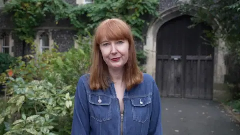 Elise Burns, who is wearing a denim shirt and has shoulder length hair, faces towards the camera.