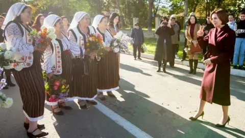 EPA Four smiling women wearing traditional Moldovan dress, including a white head-scarf, brown skirt and patterned waistcoat, welcome President Maia Sandu at a campaign event in Telenesti, 92km north of Chisinau, Moldova. The women are holding flowers, and the president makes a 'v for victory' sign with her hand.