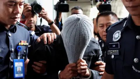 Reuters A man linked to Global Ikhwan Service and Business Holdings (GISB) covers his face as he is escorted by police officers after being charged in a court in Putrajaya, Malaysia September 18, 2024.