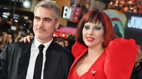 Getty Images Actor Joaquin Phoenix wearing a black suit and tie and white shirt on a red carpet with Lada GaGa