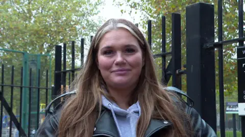 Danielle O'Leary with long hair cascading over her shoulders standing in front of a black gate with trees in the background