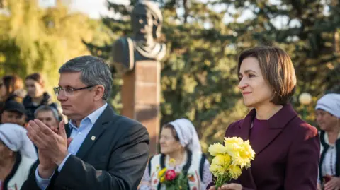 Matthew Goddard President Maia Sandu holding a small bunch of flowers during a campaign visit. She is wearing a purple suit jacket. A man standing on the left is applauding. There are people dressed in traditional clothing in the background.