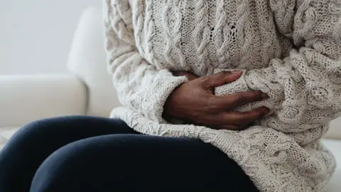 Getty Images Stock photo of a young woman wearing a heavy wool junper places her hands over her abdomen while sitting on a sofa at home.