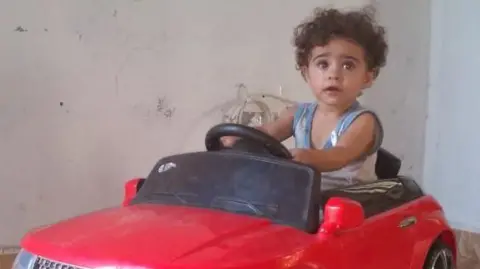 Family handout A young child sits in a large, red toy car