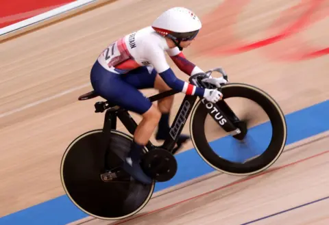 Getty Images Laura Kenny wears a blue and white team GB lycra bodysuit and white helmet, clipped into a black racing bike as she hurtles round a velodrome track.