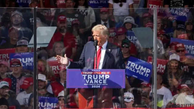 Trump speaks at a 'Trump-Vance' podium in North Carolina behind a glass protector. Behind him, supporters hold up 'Trump Will Fix It' signs