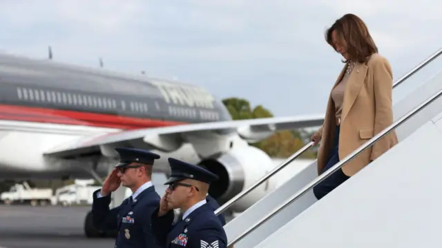 Kamala Harris exiting her plane in North Carolina, wearing a beige coat. In the background is Trump's plane, also parked on the tarmac