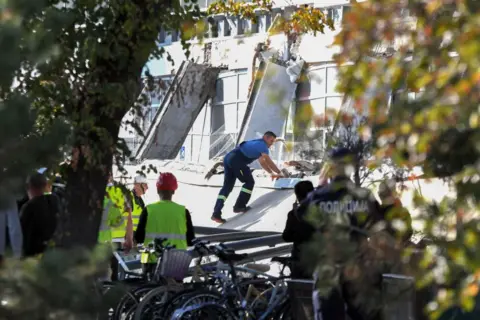 AFP via Getty Images A rescuer leans into and appears to pull on a piece of concrete that collapsed outside a railway station in Serbia while rescuers in hard hats and police officers look on