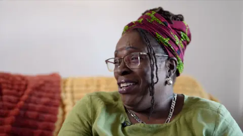 Jean, a woman wearing a light green top, with glasses and her hair wrapped in a purple and green scarf, and a few braids hanging loosely at the front, sitting on a sofa and smiling as she looks at someone to the left of the camera.