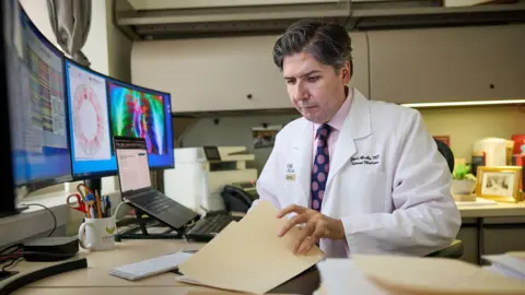 Matt Miller/WashU Medicine Man in a pink shirt and tie with a white lab coat flips through medical reports in his office.