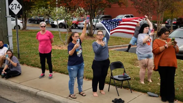 People standing on a sidewalk with their phones out, waving an American flag while capturing Donald Trump's motorcade drive by