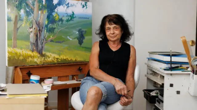 June Carey, 70, from Chico, California sat on chair with landscape painting behind her