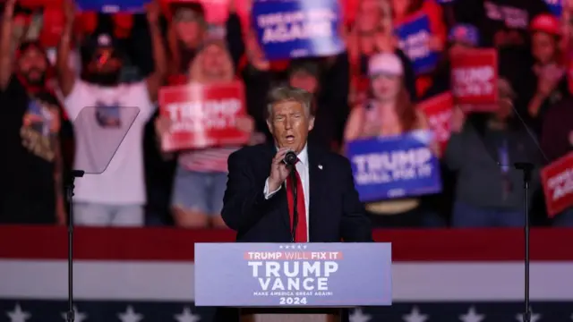 Donald Trump speaks at a podium at a rally in Greensboro. He holds the mic, and is wearing a blue suit with a red tie.