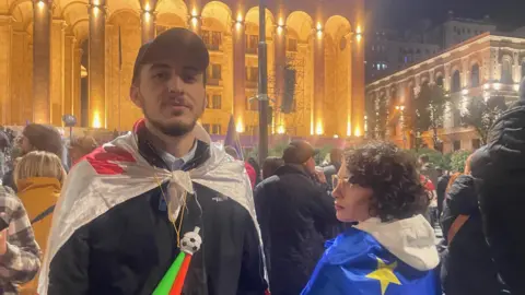 A young man draped in a Georgian flag stands next to a crowd in the street