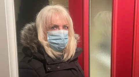 Andrea is standing at her open front door. The door is red and you can see a set of stairs behind her. She is wearing a black coat with a furry hood, and a blue face mask