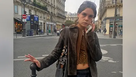 Paola Goddard A young woman with brown hair stands in the street, resting her hand on a bollard. She is wearing a brown jacket and a leopard-print top. She is holding her phone up to her face and has a serious expression