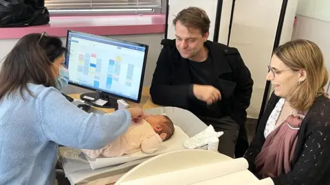 BBC A baby wearing a light pink suit is weighed by a midwife with long brown hair and wearing a light blue jumper. The baby's parents, a brown-haired man and a blonde woman wearing glasses, look on and smile